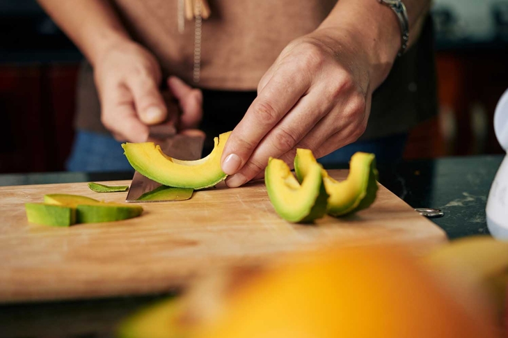 Hands peeling cut avocado slices on wooden board