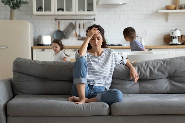 Young tired single mother suffers from headache closed eyes touch forehead sitting on couch while her daughter and son running around her and shouting, female babysitter feels exhausted by noisy kids