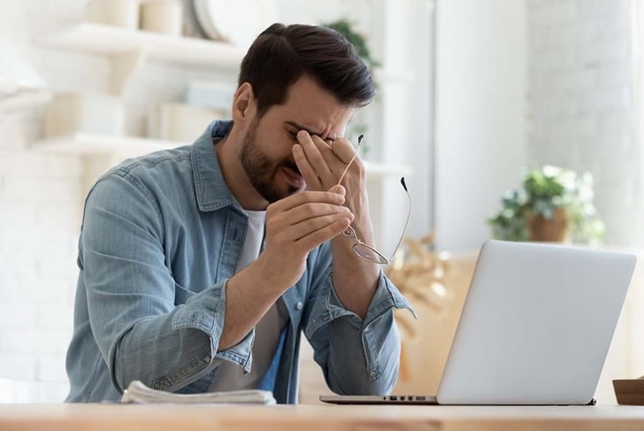 Tired young man feel pain eyestrain holding glasses rubbing dry irritated eyes fatigued from computer work, stressed man suffer from headache bad vision sight problem sit at home table using laptop